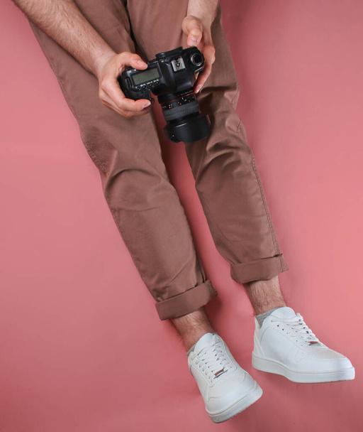 Άνδρας σε μπεζ παντελόνι και λευκά sneakers κρατώντας φωτογραφική μηχανή, ενώ κάθεται πάνω από ροζ φόντο. Στο πάνω μέρος. Φωτογραφία στούντιο - Φωτογραφία, εικόνα