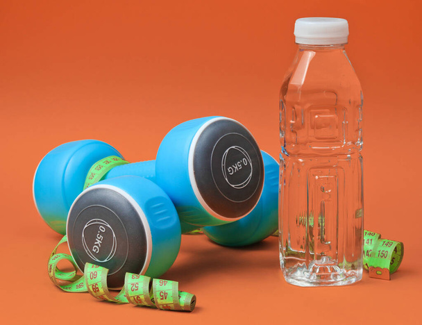 Healthy lifestyle still life. Dumbbells, ruler,  bottle of water on orange background. Slimming concept - Foto, imagen
