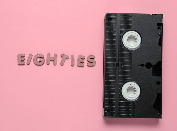 Retro style concept, 80s. Video cassette on pink pastel background with the word Eighties from wooden letters. Top view, minimalism - Photo, Image