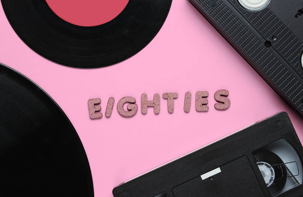 Retro stijl concept, jaren 80. Videocassette en vinylplaten op roze achtergrond met het woord jaren tachtig uit houten letters. Bovenaanzicht, minimalisme - Foto, afbeelding
