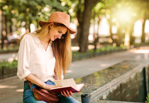 Νεαρή φοιτήτρια ντυμένη με ρούχα casual στυλ διαβάζοντας ένα βιβλίο, ενώ κάθεται σε μια πόλη σε εξωτερικούς χώρους. - Φωτογραφία, εικόνα