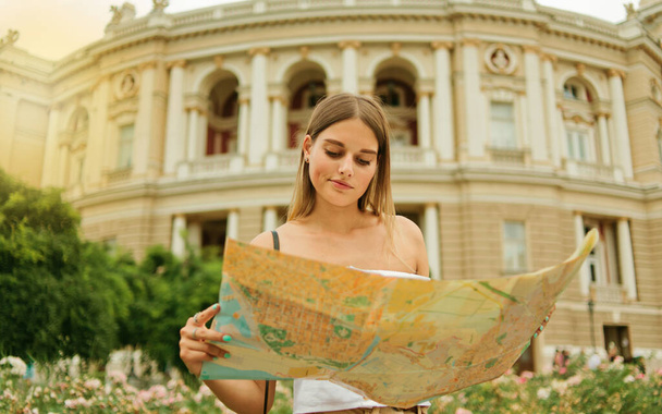Jonge, verwarde vrouwelijke toerist houdt in haar handen kaart van de stad tegen de achtergrond van de architectuur van een toeristische stad. Reizen naar nieuwe plaatsen - Foto, afbeelding