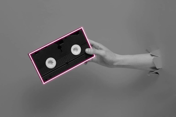 Female hand holds video cassette with neon frame through torn  paper background. - Photo, Image