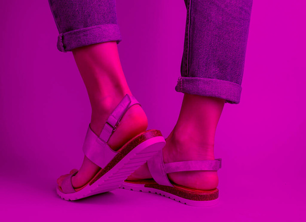 Fragment ženských nohou v modrých džínách a módních kožených sandálech. Dámské stylové letní boty. Minimalistická módní střelba. Růžové neonové světlo - Fotografie, Obrázek