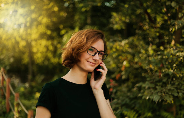 Νεαρή χίπστερ κοπέλα με γυαλιά που μιλάει στο τηλέφωνο έξω. - Φωτογραφία, εικόνα