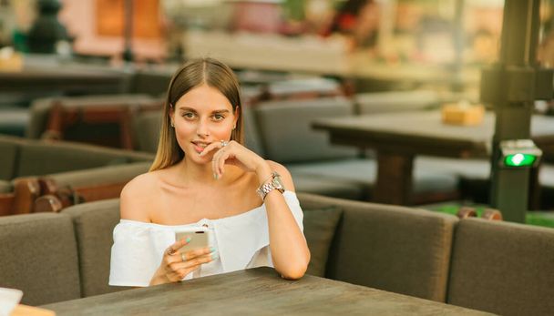 Bir sokak kafeteryasındaki masada otururken elinde akıllı telefon tutan şık bir genç kadın. - Fotoğraf, Görsel
