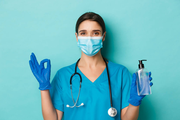 Concept of covid-19 and quarantine concept. Close-up of confident female doctor in medical mask, gloves and scrubs showing okay sign, advice to use hand sanitizer to be safe from coronavirus - Фото, зображення