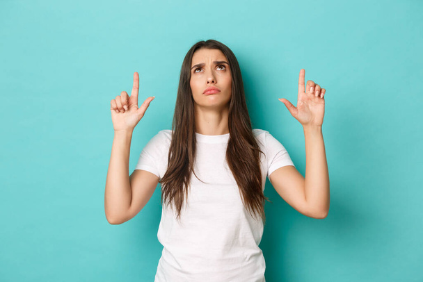 Portrait of confused brunette woman in white t-shirt, looking and pointing fingers up, cant understand something, standing over blue background - 写真・画像