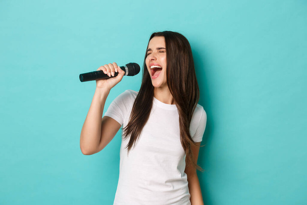 Retrato de mujer morena despreocupada, divirtiéndose en el bar de karaoke, cantando en micrófono mientras está de pie sobre fondo azul - Foto, Imagen