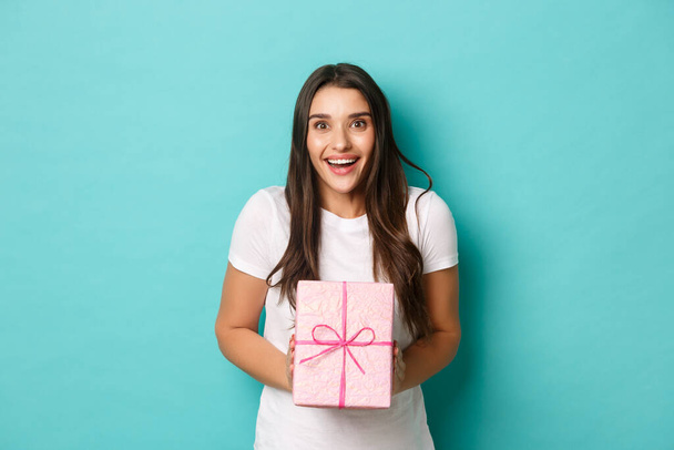 Concept of holidays and celebration. Image of excited woman looking happy, smiling and receiving gift wrapped in pink box, standing over blue background - Photo, image