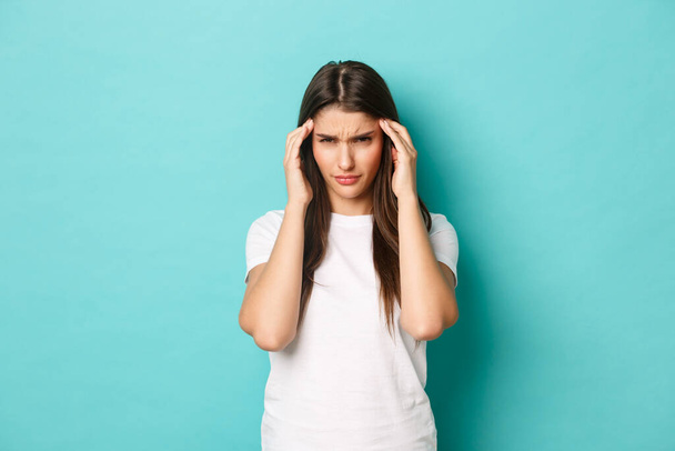 Изображение расстроенной молодой женщины, пытающейся сосредоточиться, трогающей голову и щурящейся, страдающей головной болью, стоящей на синем фоне - Фото, изображение