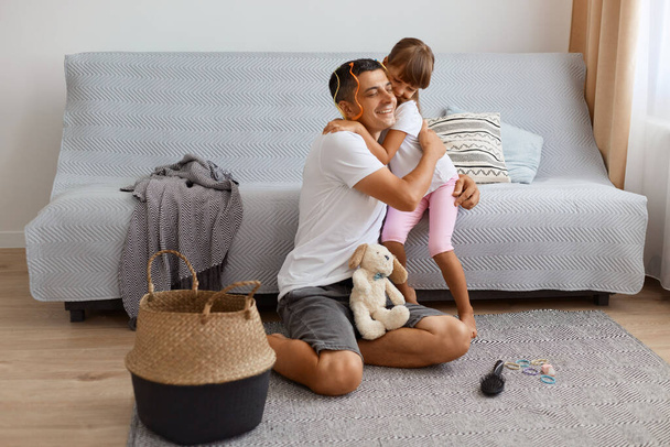 Indoor shot of happy positive young adult father spending time with his daughter while sitting on floor near sofa in living room, family hugging with love and gentle, playing together. - Φωτογραφία, εικόνα