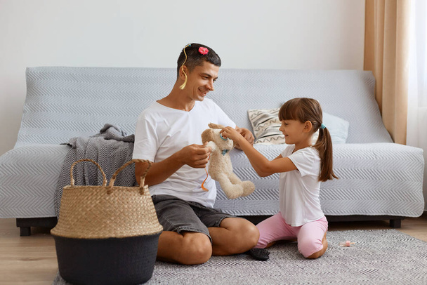 Positive young adult father spending time with his daughter while sitting on floor near sofa in living room, dad holding soft dog toy, kid making glasses for toy with scrunchies, having fun. - Photo, Image