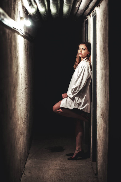Dívka v dlouhé bílé košili s depresivním výrazem v tmavé chodbě. - Fotografie, Obrázek