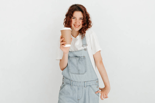 Jonge vrouw met een overall met een hete kop koffie, over een witte muur. Vrijetijdsconcept - Foto, afbeelding