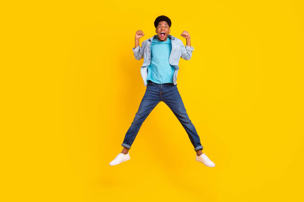 Photo of sweet lucky dark skin man wear jeans shirt jumping high rising fists smiling isolated yellow color background - Valokuva, kuva