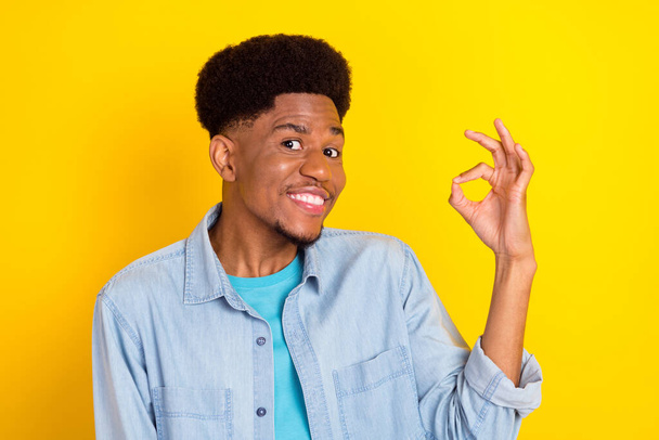 Photo of sweet cute dark skin guy dressed denim shirt smiling showing okey sign isolated yellow color background - Foto, afbeelding
