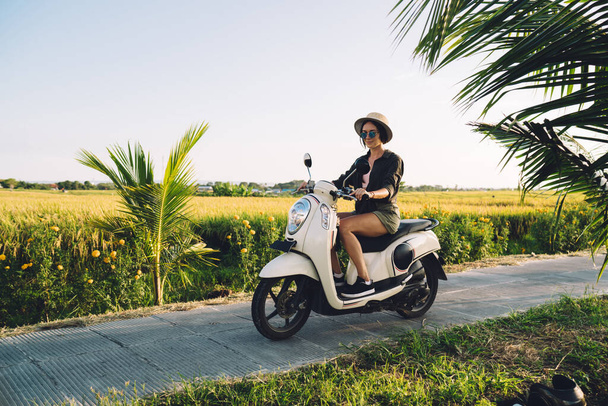 Side view of attractive Caucaisan tourist driving old fashioned scooter on travel path enjoying sunny weather at nature environment, beautiful hipster girl testing vintage moped during journey - Foto, Imagem