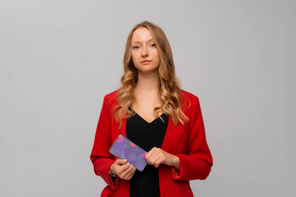 hermosa y confiada mujer de negocios sostiene un teléfono móvil en sus manos, de pie en blazer rojo sobre fondo gris - Foto, Imagen