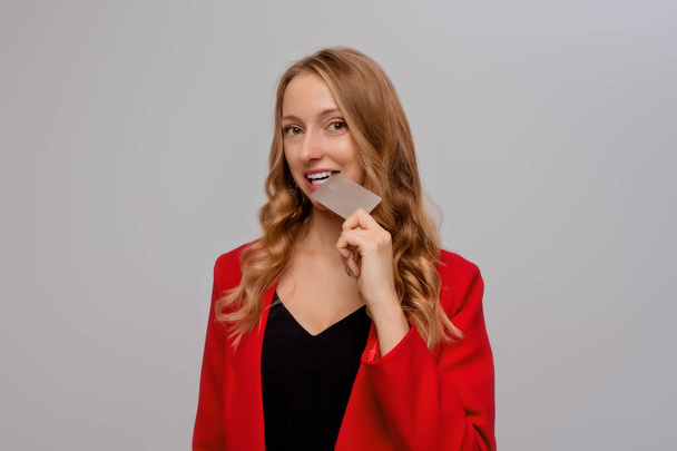 Playful young blonde woman biting credit card, thinking of doing online shopping, standing in red blazer against gray background - Foto, immagini