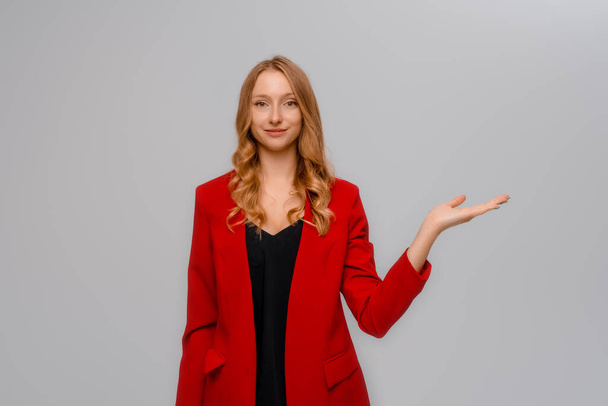 Beautiful young blonde woman with open hand palm, standing in red blazer against gray background - Foto, Bild