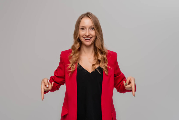 Smiling excited blonde young woman pointing fingers to the down, looking happy, showing to the advertisement, demonstrating promo offer, stands in red blazer against gray background - Zdjęcie, obraz
