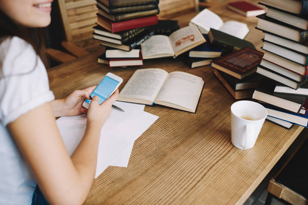 High angle of young female student surfing internet on smartphone while working at project sitting at wooden table with papers books and mug - Valokuva, kuva