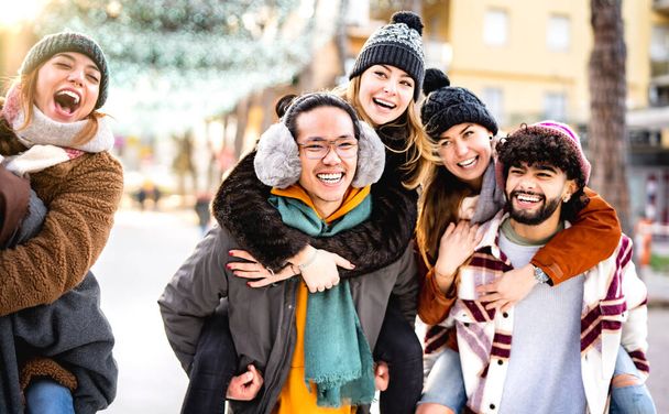 ピギーバック移動の冬の旅行の場所を歩く幸せな多文化の友人-屋外で一緒に楽しむ幸せな男と女の子との日常生活スタイルの概念-暖かいバックライトフィルター - 写真・画像
