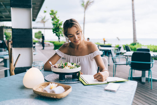 Positive young ethnic female taking notes in diary while having healthy lunch on terrace of tropical cafe against cloudy sky and ocean - Fotó, kép
