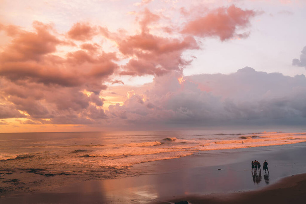 Group of people standing on wet sandy coast near waving orange sea during beautiful sunset and looking on colorful clouds - Fotó, kép