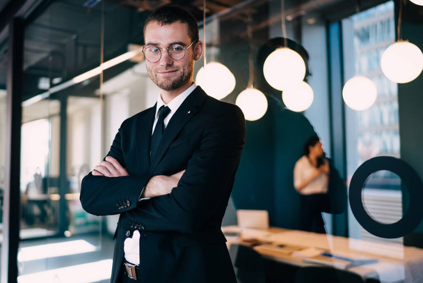 Content young businessman in formal wear and eyeglasses standing with arms folded near office glass wall and looking at camera confidently - Foto, immagini
