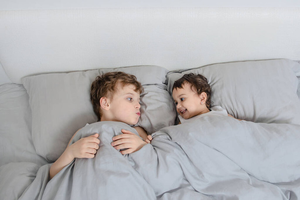Brother and sister morning concept. Little boy look to his sister girl in the bed and smiles to each other after waking up. They are wearing white t-shirts - Zdjęcie, obraz