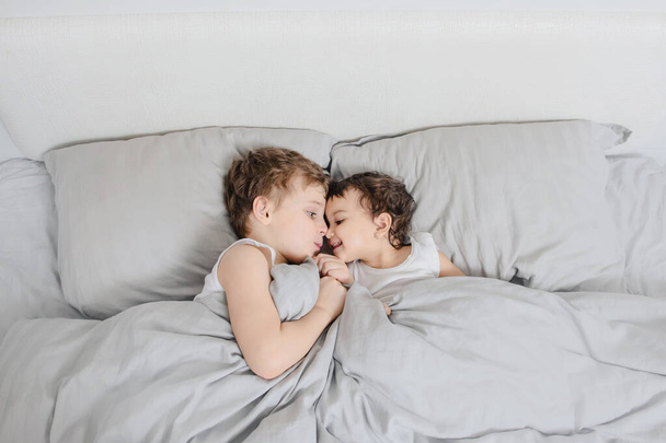 Brother and sister morning concept. Little boy look to his sister girl in the bed and smiles to each other after waking up. They are wearing white t-shirts - Fotó, kép