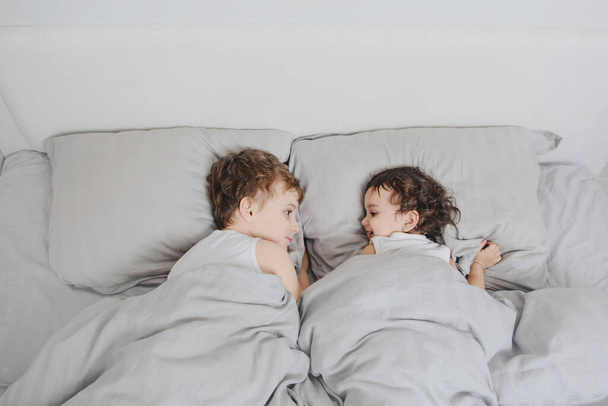 Erkek ve kız kardeş sabah kavramı. Küçük çocuk yatakta kız kardeşine bakar ve uyandıktan sonra birbirinize gülümsersiniz. Beyaz tişört giyiyorlar. - Fotoğraf, Görsel
