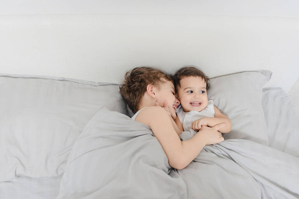 Broer en zus ochtend concept. Kleine jongen knuffelt zijn zus meisje in het bed en lacht na het wakker worden. Ze dragen witte t-shirts. - Foto, afbeelding