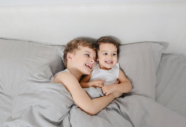 Poranna koncepcja brata i siostry. Mały chłopiec przytulający siostrę do łóżka i śmiejący się po przebudzeniu. Noszą białe koszulki. - Zdjęcie, obraz