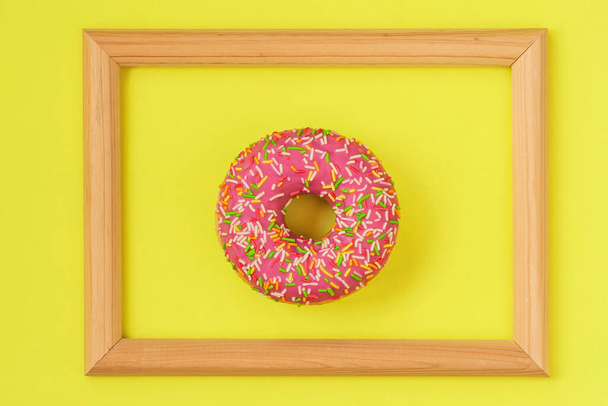 Fresch bright glazed donut in wooden frame on yellow background, unusual abstract sweet art - Foto, Imagem