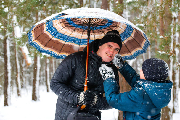 Junger Mann und lustiger Junge auf ländlichem, schneebedecktem Hintergrund, Regenschirm haltend und kämpfend. Glückliche Familie, kaltes Wetter. Lebensstil. Verschneiter Winter, verloren im Wald. Älterer Bruder. Lächelnd und scherzend. - Foto, Bild