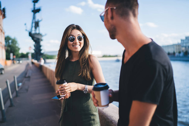 Young glad female in casual jumpsuit and sunglasses standing with smartphone and cup of coffee next to boyfriend against river and blue sky and looking at each other - Φωτογραφία, εικόνα