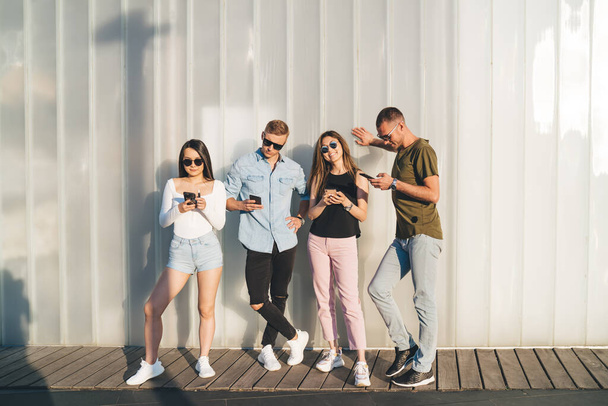 Full body group of confident multiracial friends in casual clothes and sunglasses standing near white wall and browsing smartphones together - Fotó, kép