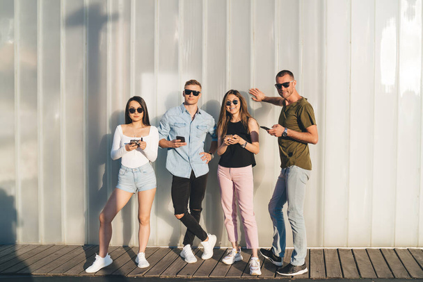 Full length portrait of trendy dressed hipster guys with cellphone gadgets posing at publicity area, youthful male and female best friends 20s using smartphone during leisure pastime in city - Foto, afbeelding