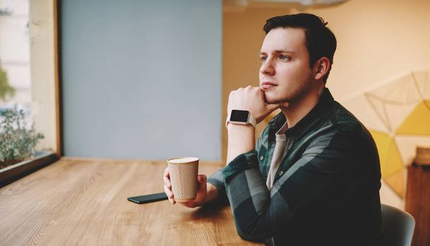 Side view of young pensive man in casual wear and smartwatch drinking hot beverage at table and thinking looking away in cafeteria - Fotó, kép