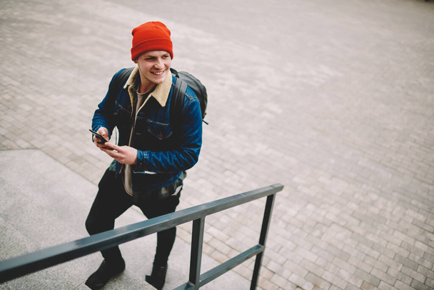 From above of male in casual clothes with backpack looking away going up stairs and surfing smartphone while walking having free time - Photo, Image