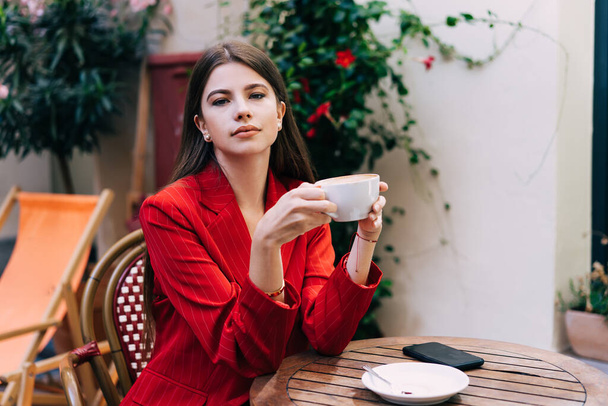 Concentrated young female sitting with cup of coffee in hands at street cafe with amazing plants by wall and looking at camera - 写真・画像