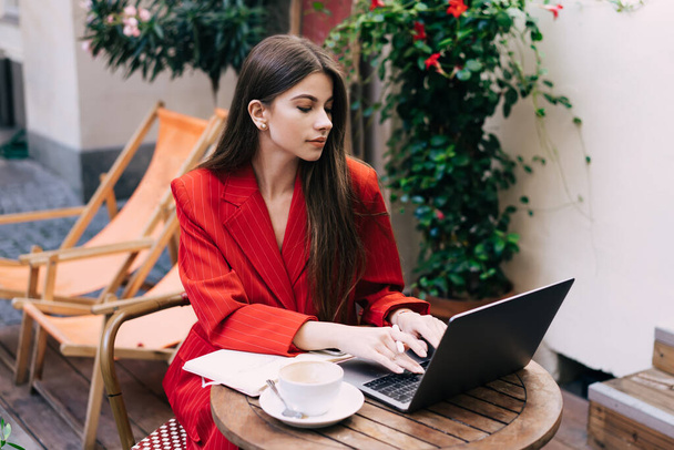 Focused young businesswoman in elegant wear typing on keyboard on laptop and looking at screen while working with coffee and notebook in outside cafeteria - 写真・画像