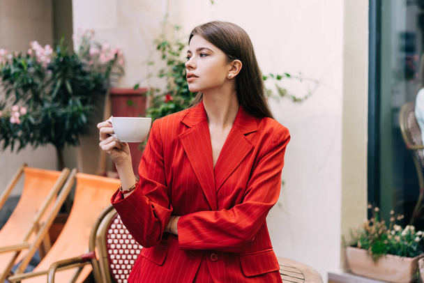 Pensive young female in stylish suit drinking cup of hot drink while standing on terrace in street cafe near table - Foto, immagini
