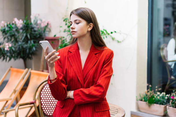 Serious young female in stylish outfit browsing mobile phone while standing near table on terrace in street cafe on daytime - Фото, изображение