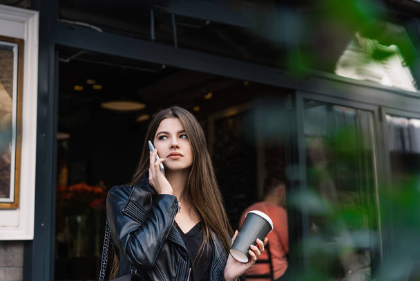 Vakava nuori nainen rento vaatteita kuppi kuumaa juomaa vastaamalla puhelun ja katselee pois seistessään kahvilan ulkopuolella - Valokuva, kuva