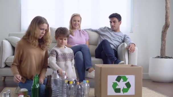 Happy caucasian family separating trash for further recycling at home. Older sister and brother putting empty plastic bottles into recycle box for ecologically friendly. Saving environment concept. - Footage, Video