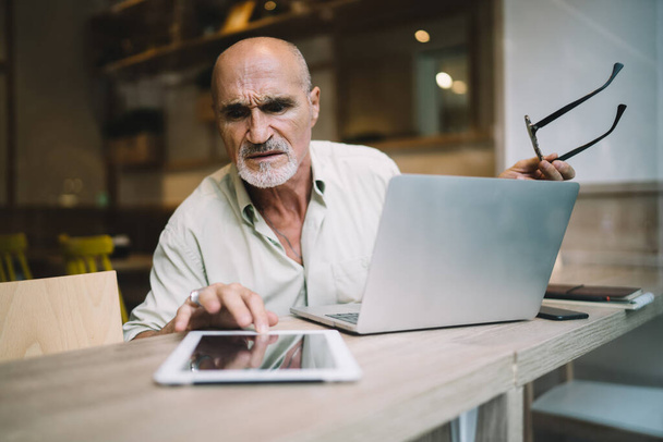 Puzzled male freelancer 60 years old confused with received gadget notification during distance working on laptop, skilled aged man checking email message on modern digital tablet using 4g internet - Foto, Imagem